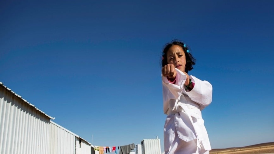 En un campamento de refugiados, una niña que lleva puesto un uniforme blanco de karateka hace un movimiento de karate frente a la cámara