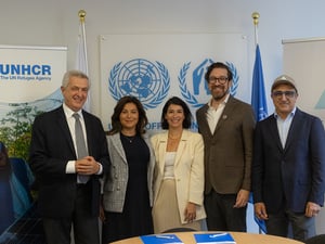En una hilera, cinco personas —tres hombres y dos mujeres— sonríen a la cámara; al fondo, banners con los logotipos de la ONU y de ACNUR.