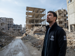 Un hombre que lleva puesta una chaqueta con el logo de ACNUR mira los escombros en una zona urbana completamente destruida.