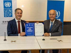 Dos hombres sonrientes que están sentados a una mesa sostienen un documento firmado en el que se lee “Acuerdo de colaboración conjunta entre el PNUD y ACNUR: 2026-2030”. Detrás de ellos se ven pancartas del PNUD y de ACNUR