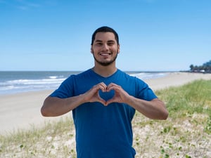 Un joven posa para la cámara en una playa haciendo el gesto del corazón con las manos.