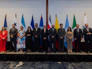 Siete hombre y tres mujeres con vestimenta formal posan de pie frente a las 10 banderas de los países y organizaciones que representan