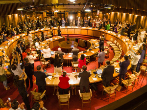 Panoraminc view of the conference room while delegates stand and clap their hands.