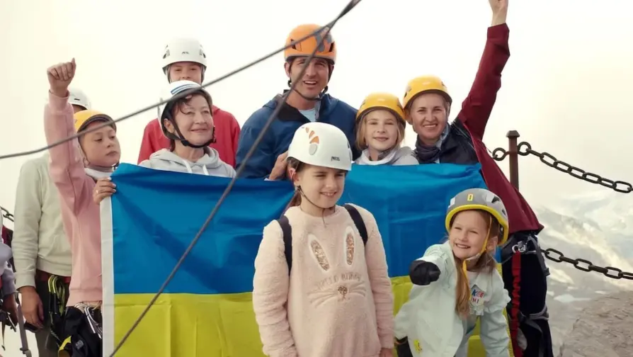 Imagem do documentário Camp Courage (2023), de Max Lowe. Uma menina refugiada da guerra na Ucrânia aparece com um capacete, vestida de rosa, em frente à bandeira da Ucrânia.