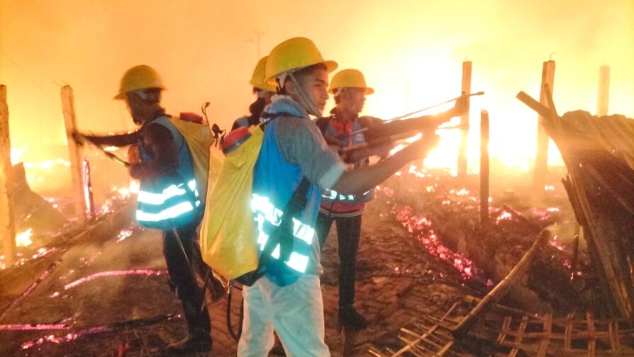 Três homens, refugiados voluntários, usando capacetes e mochilas amarelas e coletes azuis, atuam no combate ao incêndio em campo de refugiados em Bangladesh. Eles estão em meio a estruturas danificadas, tomadas pelo fogo.