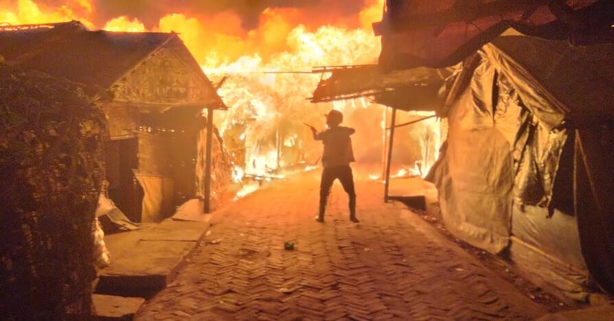 Silhueta de refugiado voluntário, atuando contra incêndio, em meio a chamas intensas, que tomam toda a imagem. Aos lados, estruturas de abrigos aparecem danificadas pelo fogo.