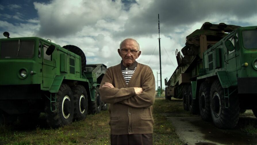 Imagem do documentário Iván (2011), de Guto Pasko. O refugiado ucraniano Iván aparece com os braços cruzados em um campo de concentração nazista na Alemanha.
