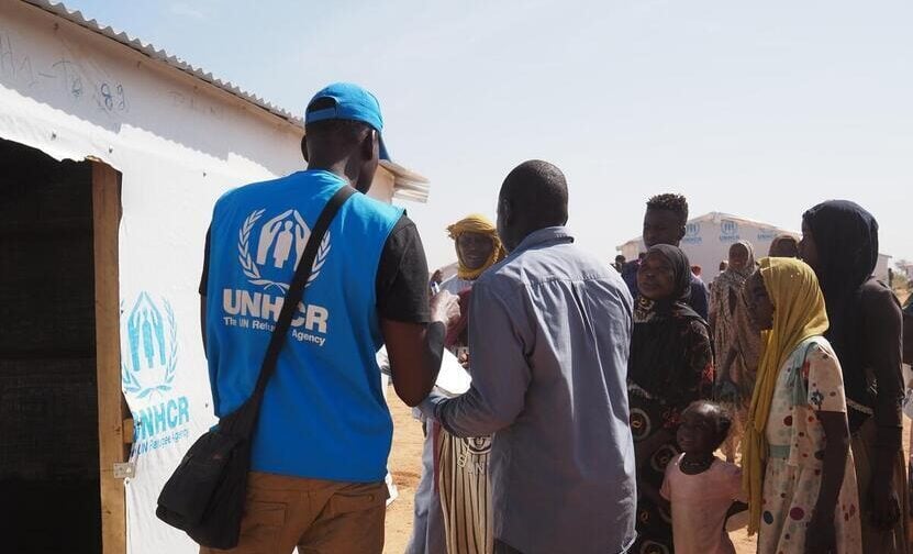 Um funcionários do ACNUR aparece alocando pessoas refugiadas sudanesas em um novo abrigo. Ele está de costas, vestindo um colete azul com o logo branco do ACNUR. Situado em frente a uma das estruturas do abrigo, de madeira, coberta com um forro branco e o logo do ACNUR azul, o trabalhador aponta com uma caneta para um documento impresso que um homem ao seu lado segura. De frente para o funcionário, pessoas refugiadas o observam atentamente. São mulheres e homens trajados com lenços na cabeça — hijab e keffiyeh, respectivamente. Entre eles, há uma criança: uma menina vestida de rosa, com o cabelo preso. Na paisagem, é possível ver, no fundo da imagem, parte de outra estrutura do abrigo, semelhante à descrita inicialmente.