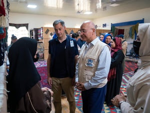 Dos hombres con vestimenta de ACNUR conversan con dos mujeres en el interior de un salón con varios tapetes coloridos sobre el suelo