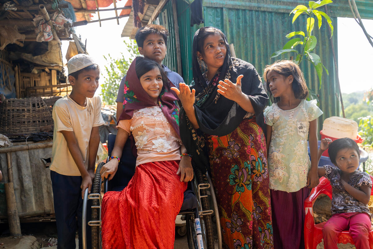 Una familias rohingya: al centro, una niña en una silla de ruedas; detrás suyo, un chico de pie; a la derecha, una mujer y dos niñas más; a la izquierda, un niño que ve hacia el frente con curiosidad.