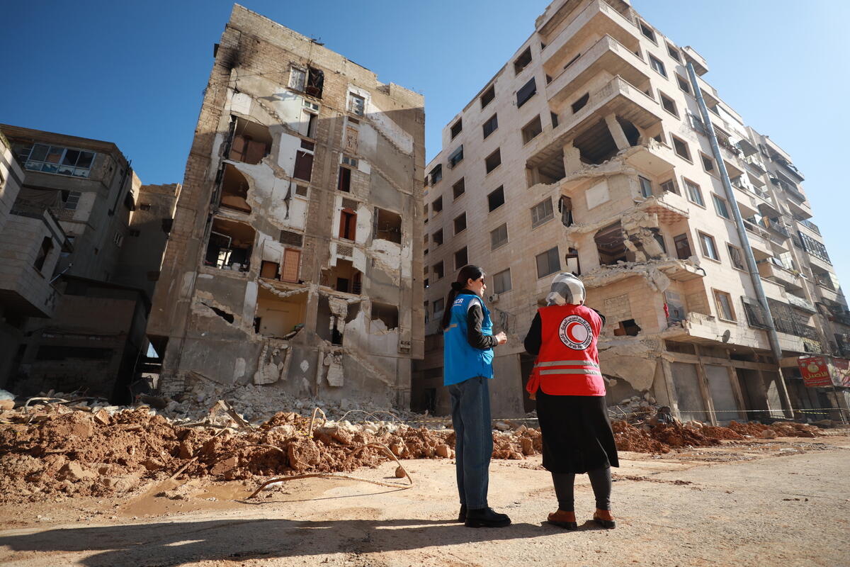 A UNHCR staff member and Red Cross staff member stand in front of a destroyed building.