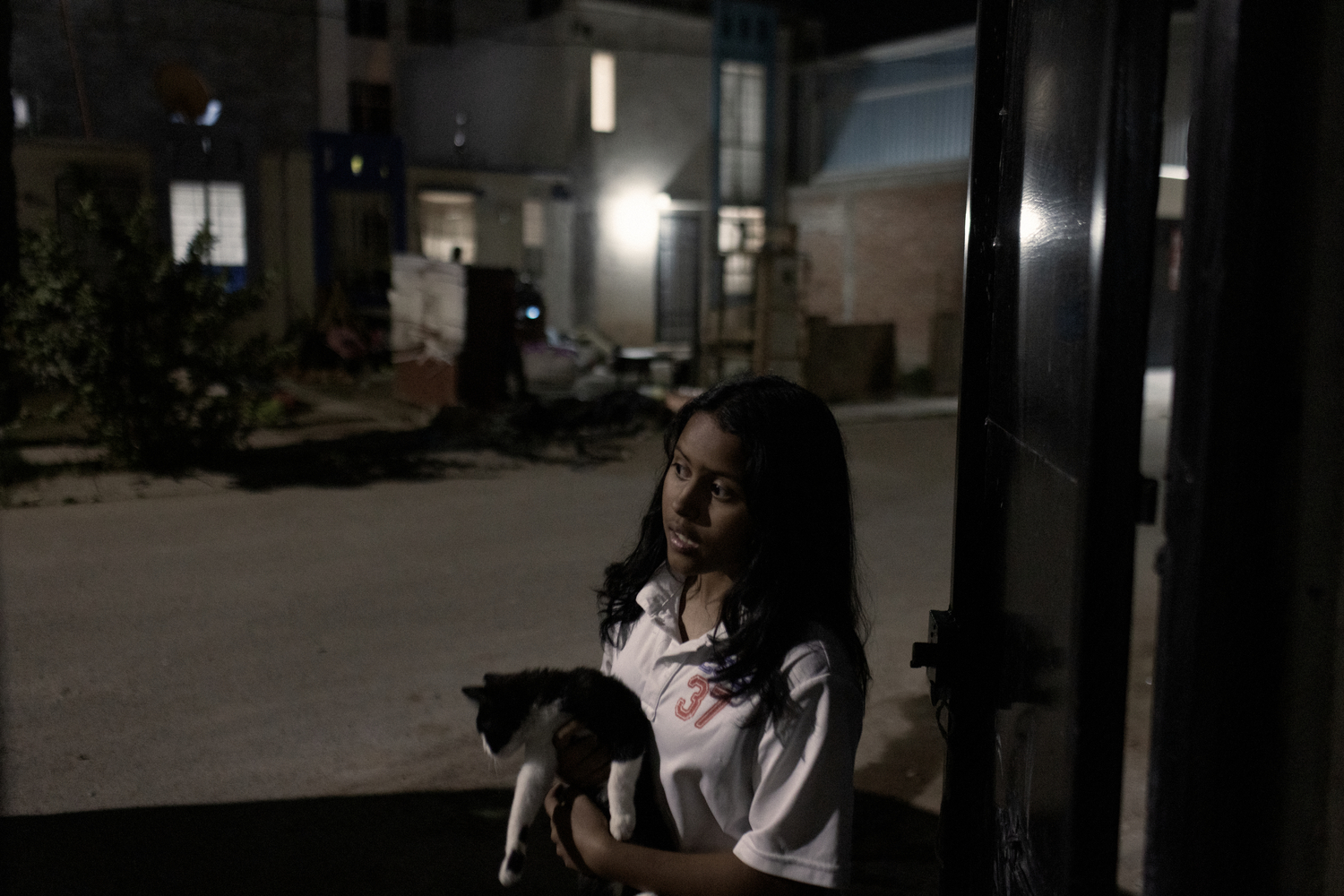 A girl stands by a gate in a dark street holding a cat. Una niña está junto a una entrada en una calle oscura con un gato en brazos.