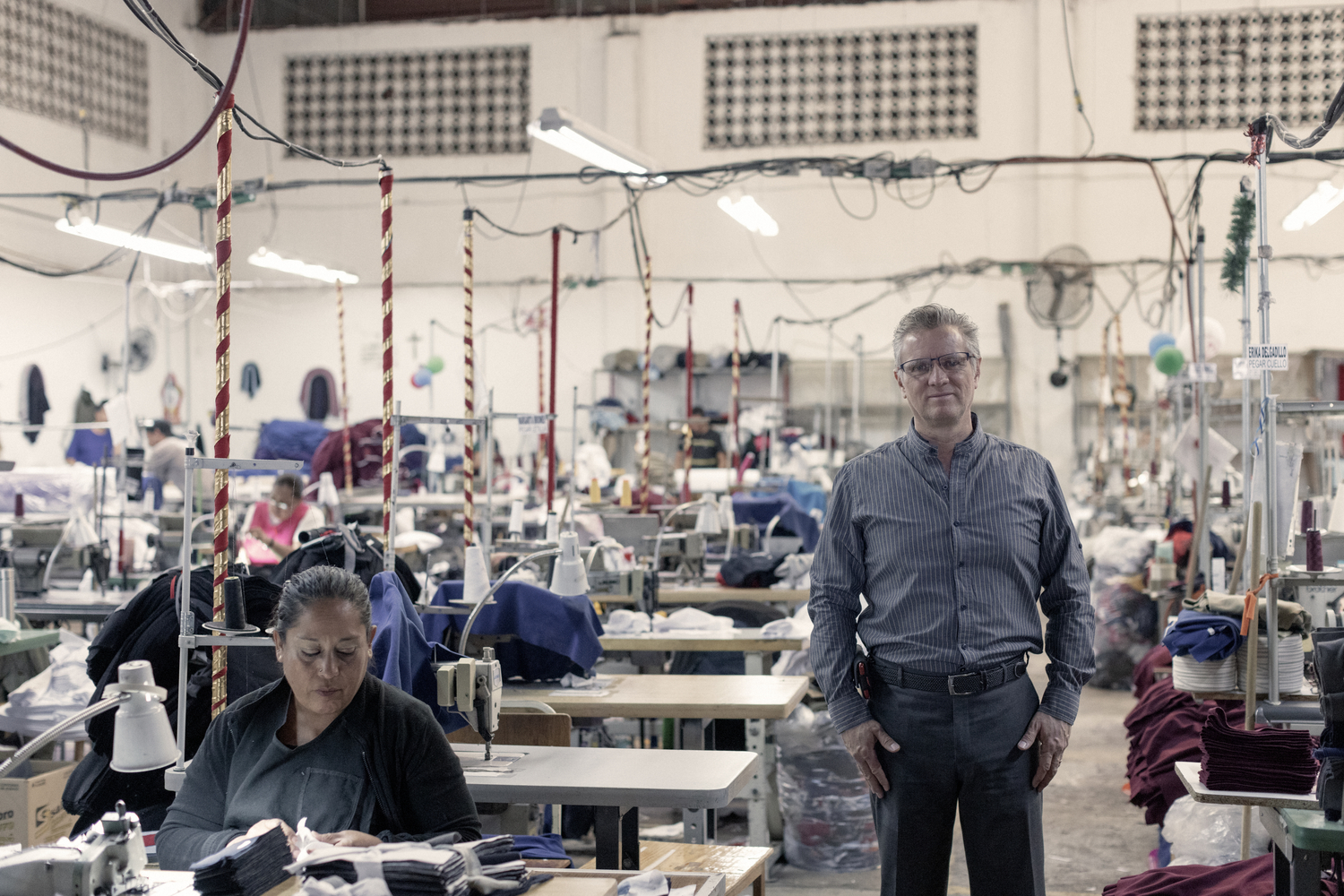 A man stands on a factory floor where women are at work using sewing machines. Un hombre en una fábrica en la que trabajan mujeres con máquinas de coser.