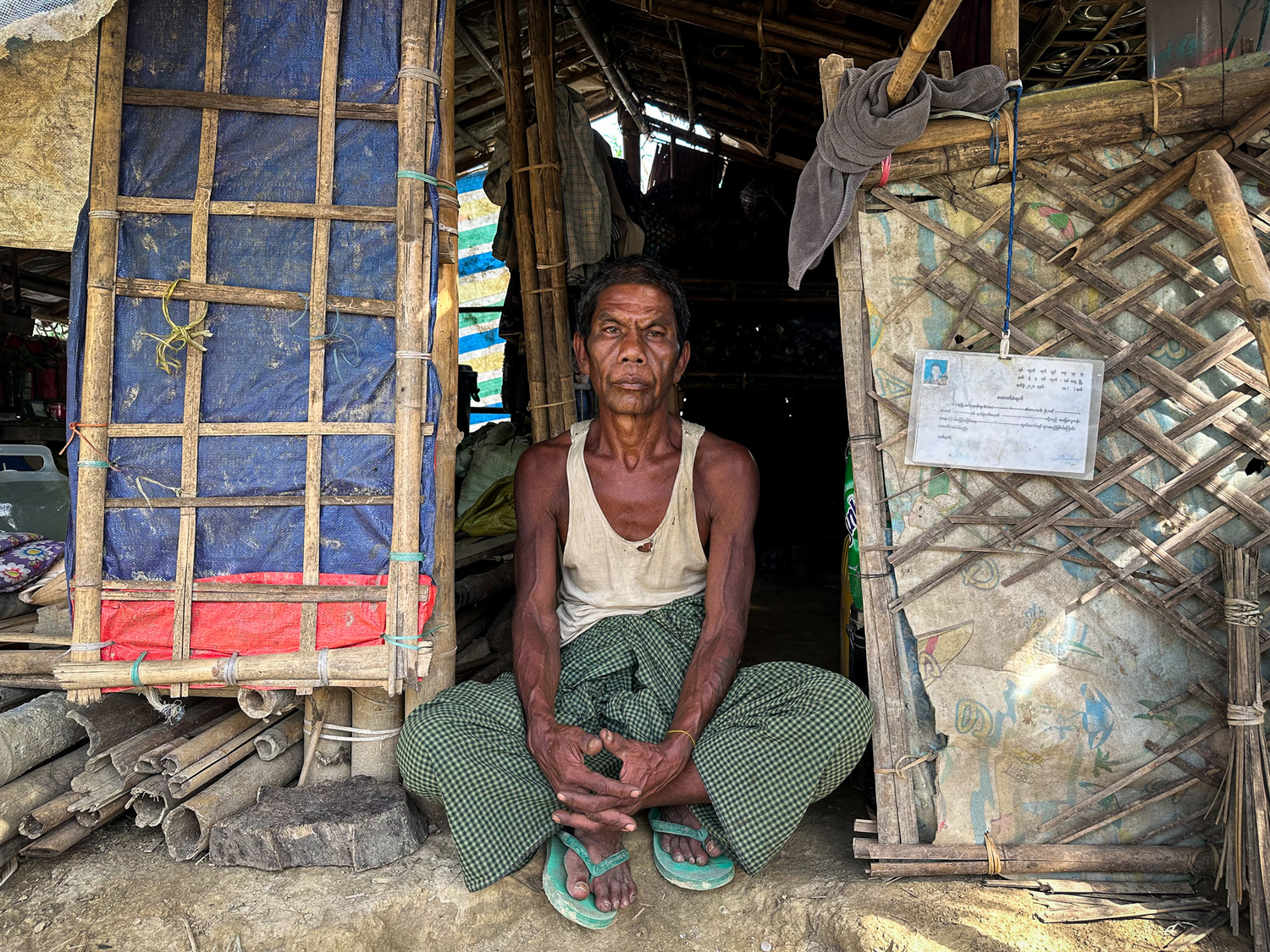 Un hombre del pueblo rakhine está sentado, con las piernas cruzadas, frente a un albergue hecho de bambú.
