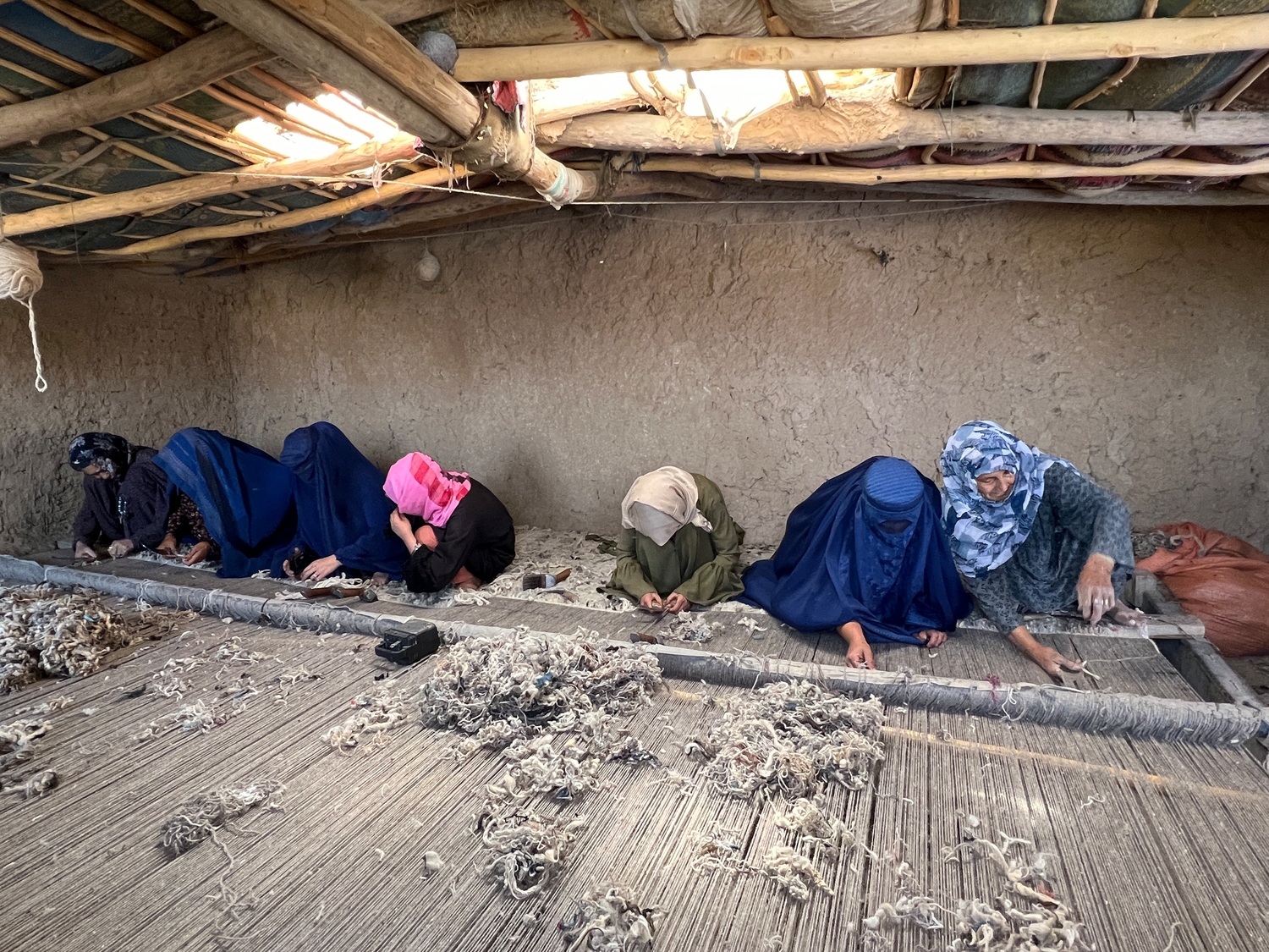 Siete mujeres están inclinadas sobre un telar gigante en el suelo de una habitación de adobe con grandes agujeros en el techo.