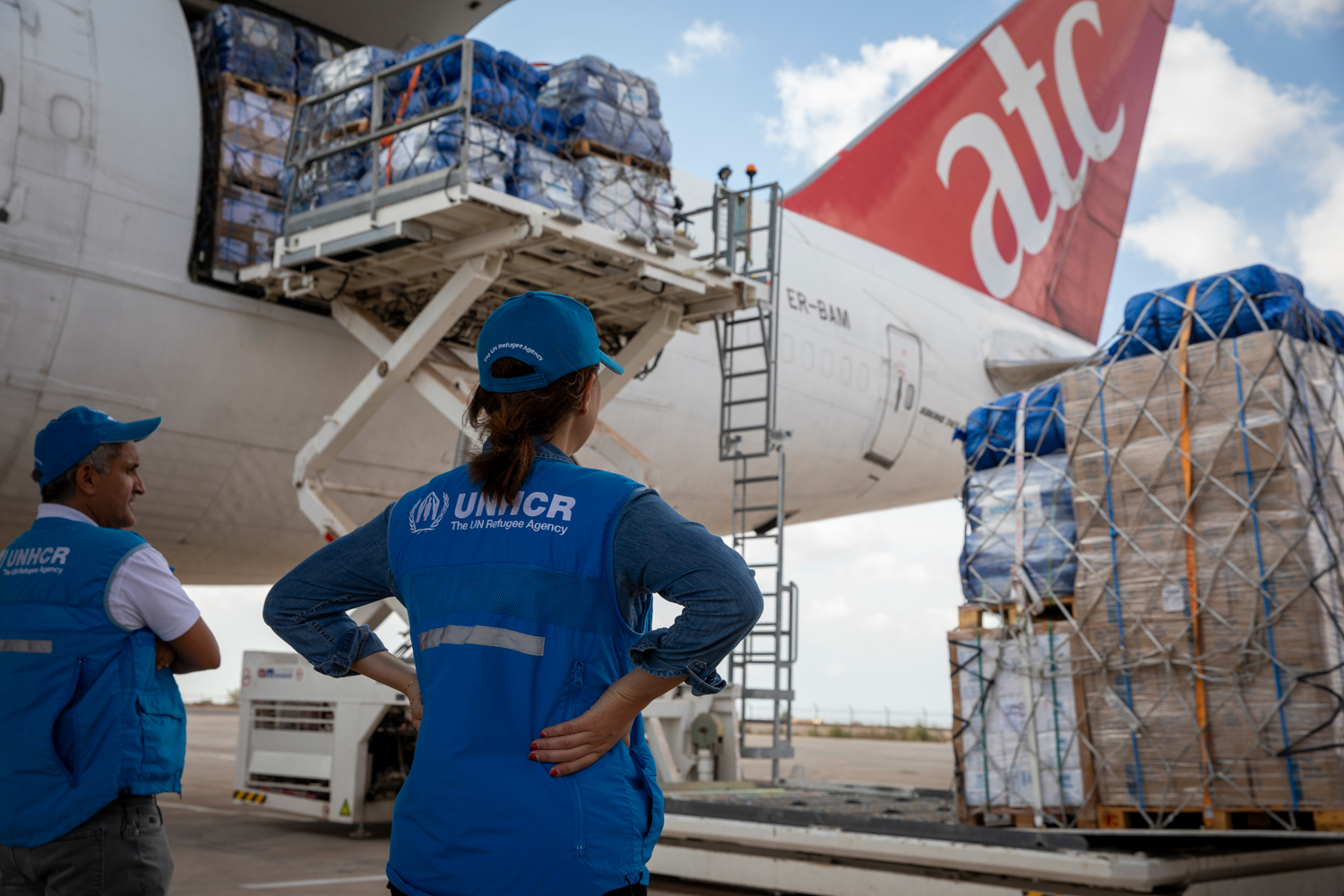 Un hombre y una mujer que llevan puestos chalecos con el logo de ACNUR observan cómo se suben suministros a una aeronave
