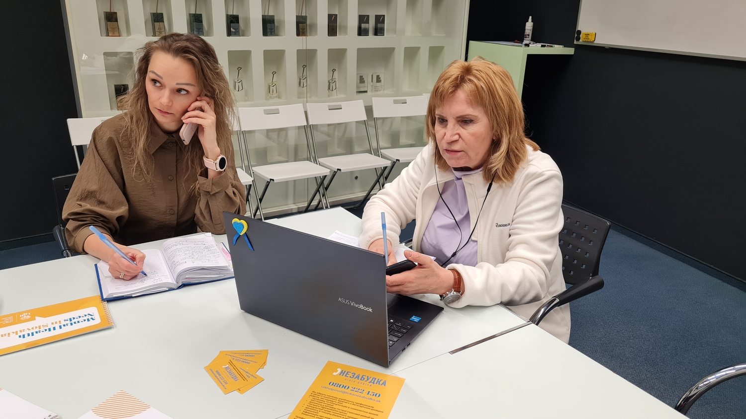 Una mujer que tiene audífonos puestos utiliza una laptop; a lado suyo está otra mujer hablando por teléfono