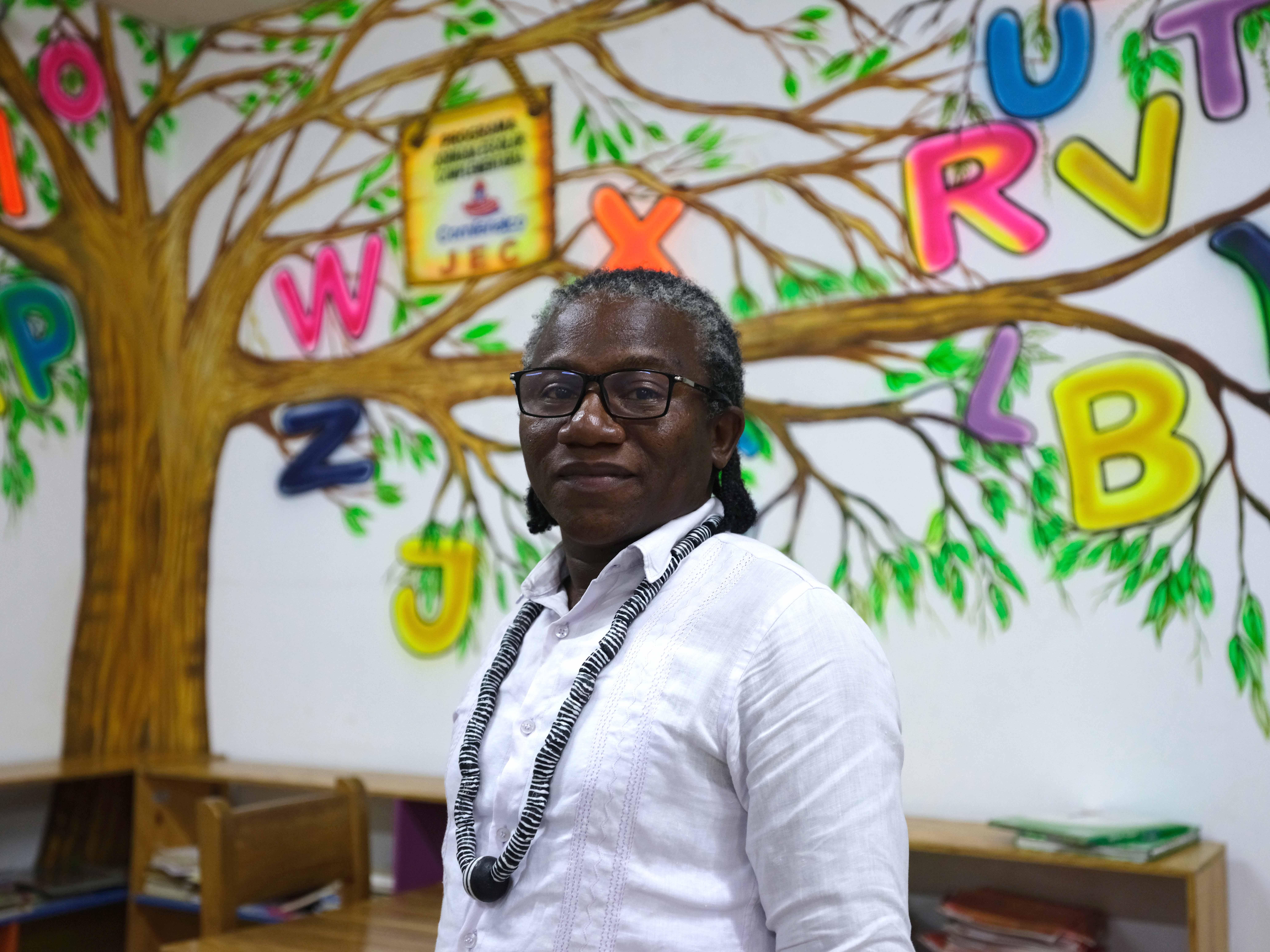 Un hombre que viste una camisa blanca manga larga y anteojos, mira hacia la cámara. Al fondo, el abecedario de colores en un salón de clases