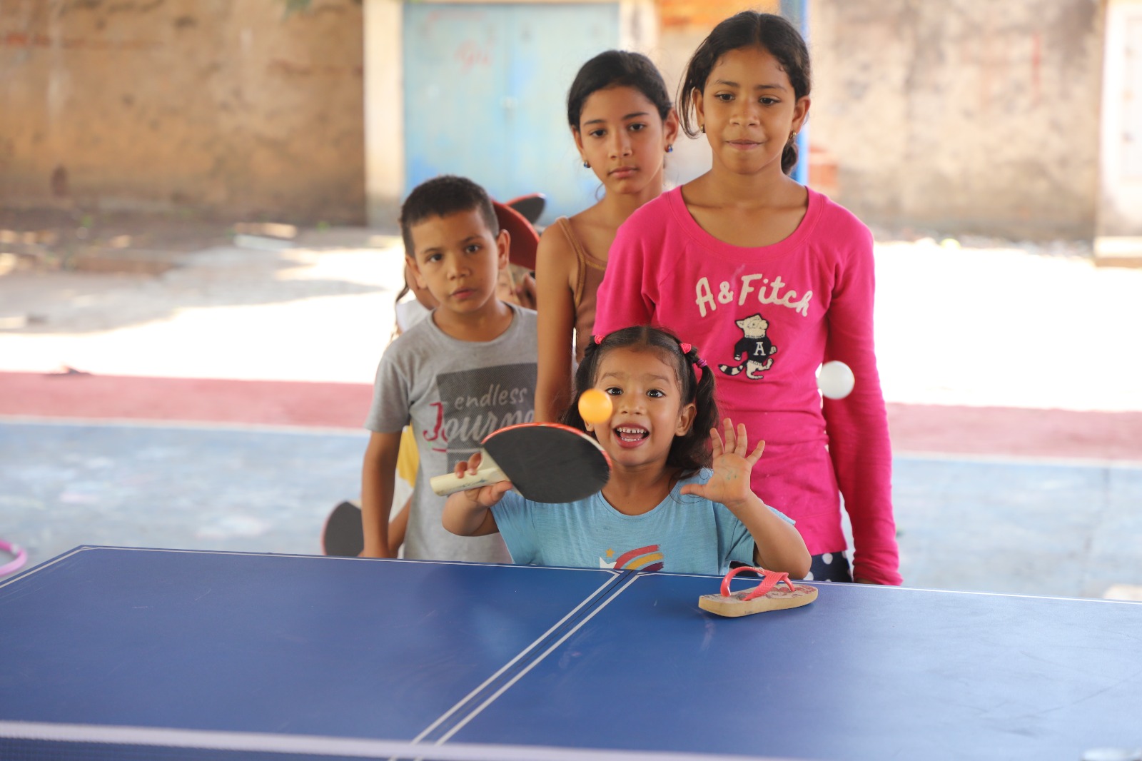 Tres niñas y un niño esperan su turno para golpear la pelota con una raqueta sobre una mesa de ping-pong color azul