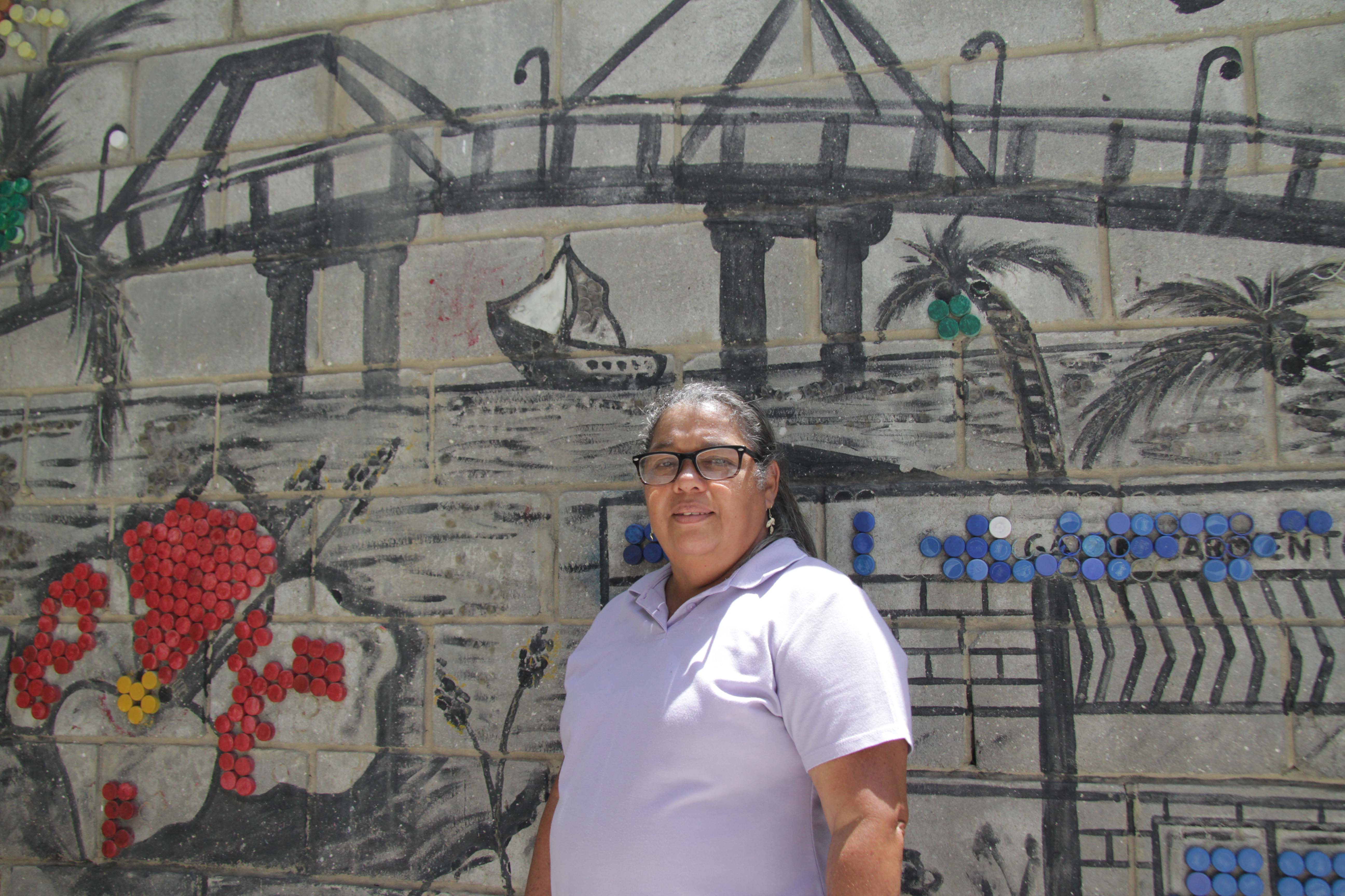 Una mujer con una camisa tipo polo color lila, frente a un mural de un puente que pasa por encima del mar.