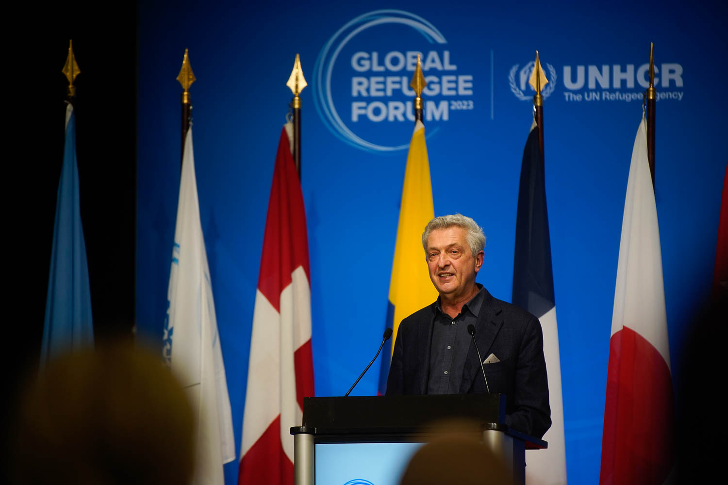 Un hombre emite un discurso desde un podio, detrás suyo, un par de banderas.