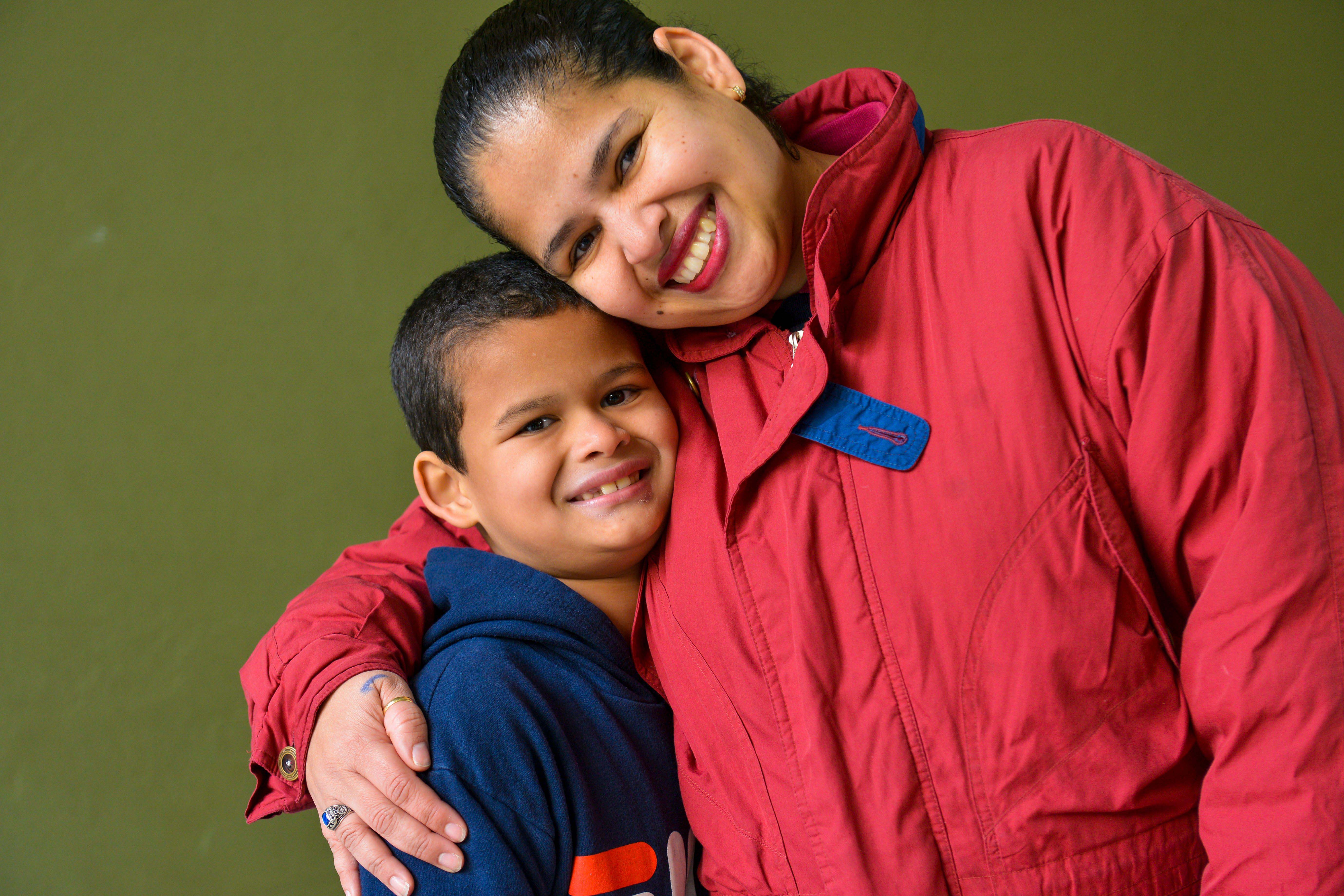 Un niño con sudadera azul marino y una mujer que viste chamarra roja, sonríen abrazados para la foto