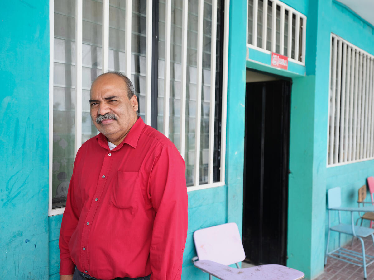Un hombre que viste camisa roja, frente a la pared azul de un aula de clases