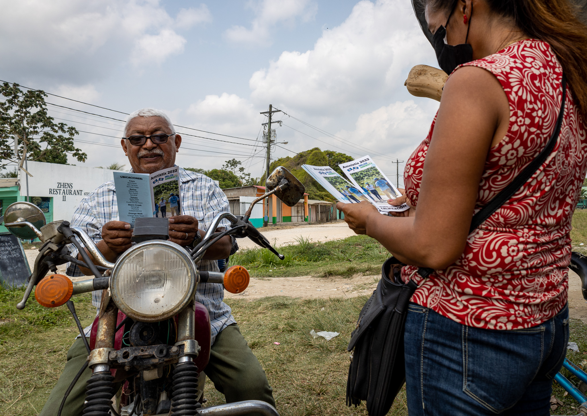 Un hombre de cabello blanco y con anteojos, lee sobre su motocicleta un folleto entregado por una mujer de blusa roja y jeans