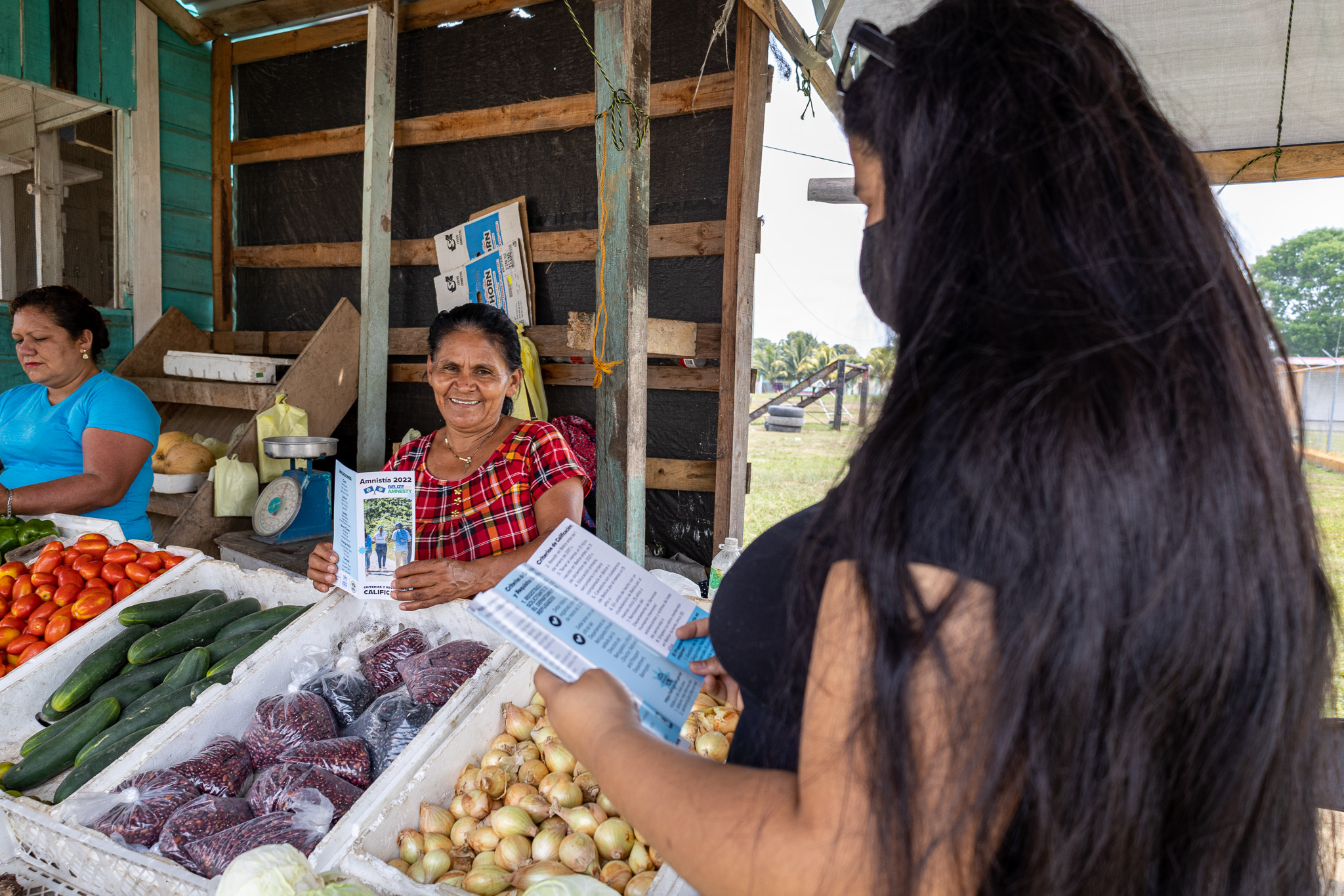 Una joven de cabello largo y oscuro entrega folletos a dos mujeres en una tienda de verduras