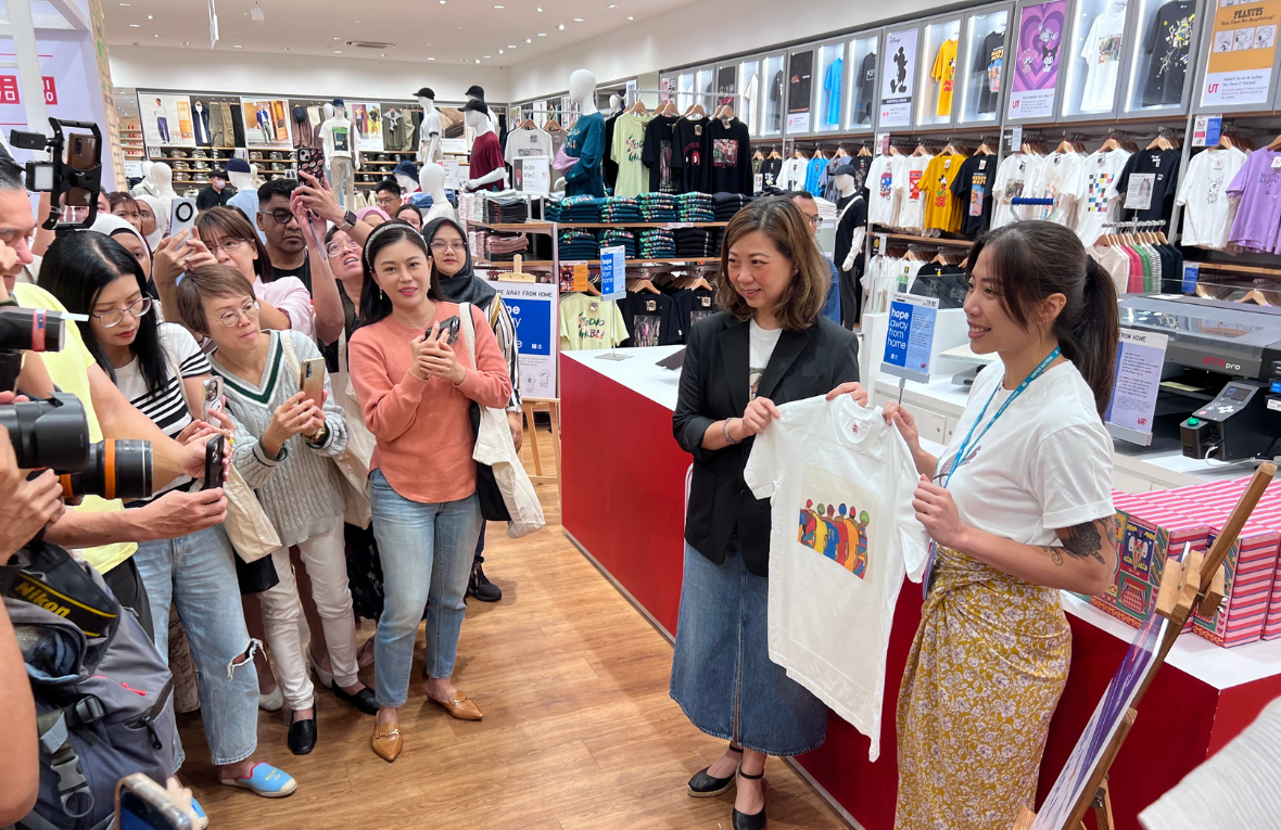 En una tienda de ropa, frente al mostrador, dos mujeres sostienen una camiseta con un estampado; frente a ellas, una multitud que toma fotografías de la prenda.
