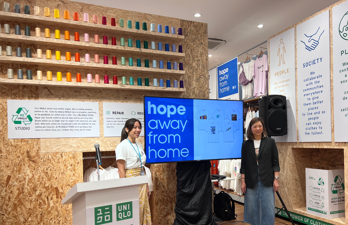 Dos mujeres junto a una pantalla que muestra una imagen de las camisetas estampadas y la leyenda &quot;Esperanza lejos del hogar&quot;, que es el lema del concurso de arte