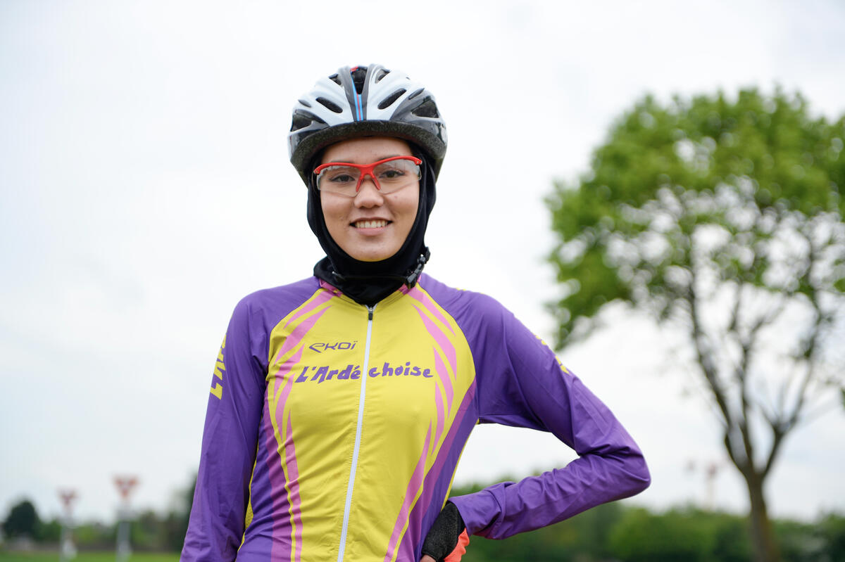 Una joven ciclista sonríe a la cámara; lleva puestas gafas y un hijab debajo del casco. Detrás suyo hay un árbol.
