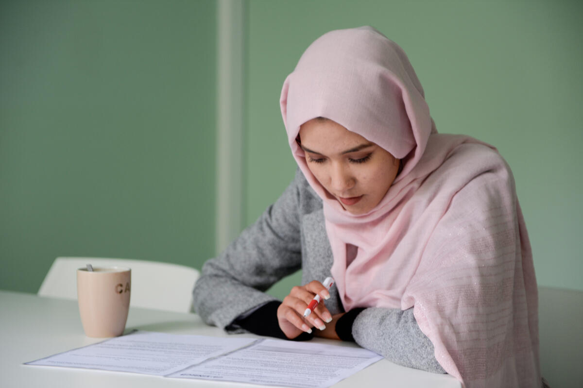 Una joven que lleva puesto un hijab color durazno está estudiando en un escritorio; junto a los papeles que tiene frente a sí hay un vaso.