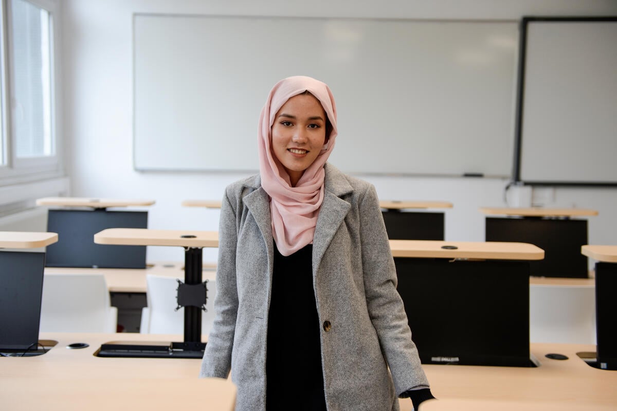 Una joven de Afganistán sonríe a la cámara desde el centro de un aula. Lleva puesto un hijab color durazno; detrás suyo, un pintarrón.