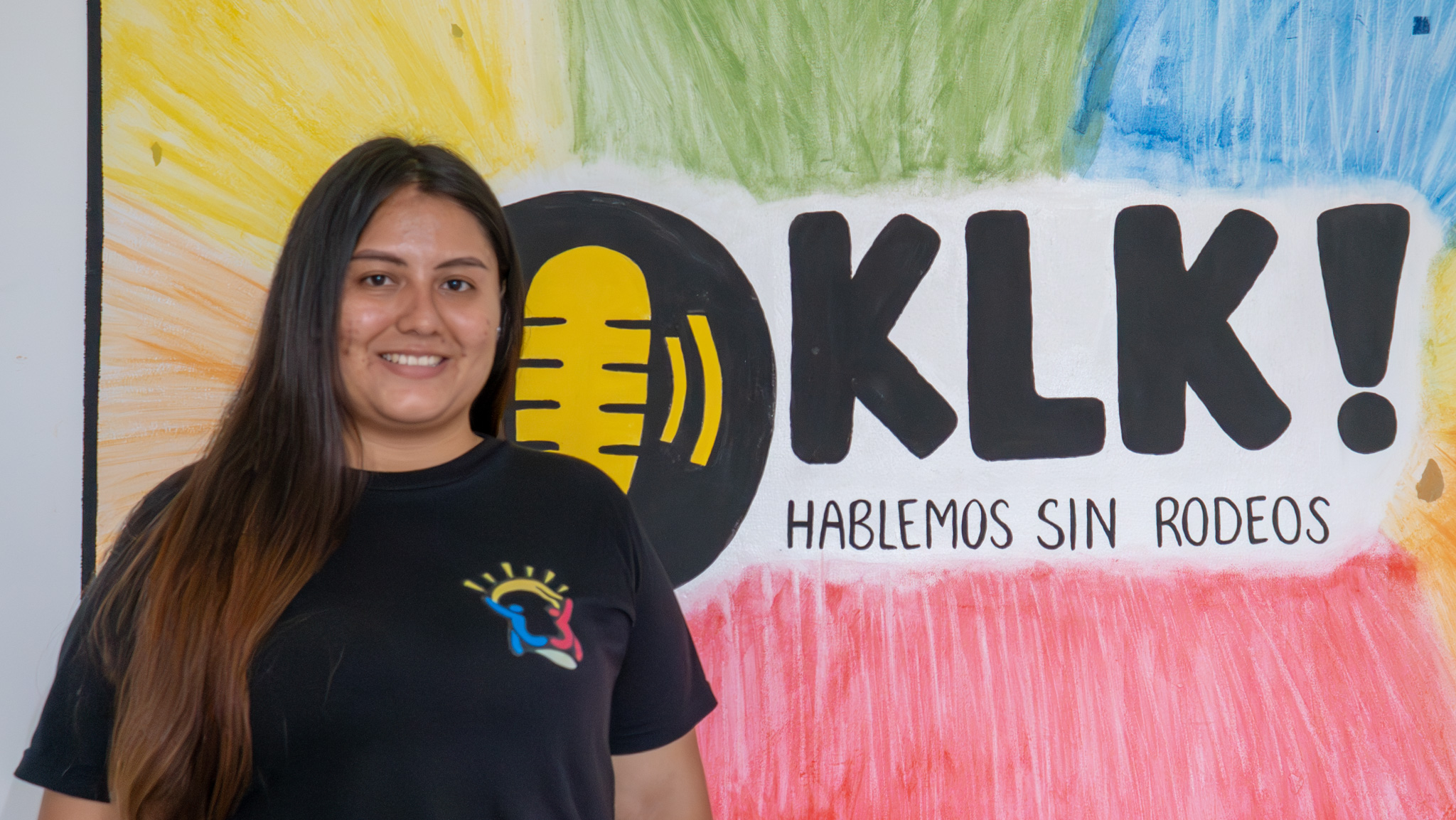 Una joven posa sonriendo para una foto delate de un mural donde se lee ¡KLK, hablemos sin rodeos!