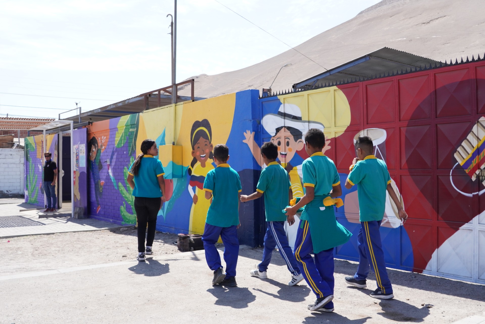 Cuatro niños y una niña caminan al lado de un mural colorido en una escuela
