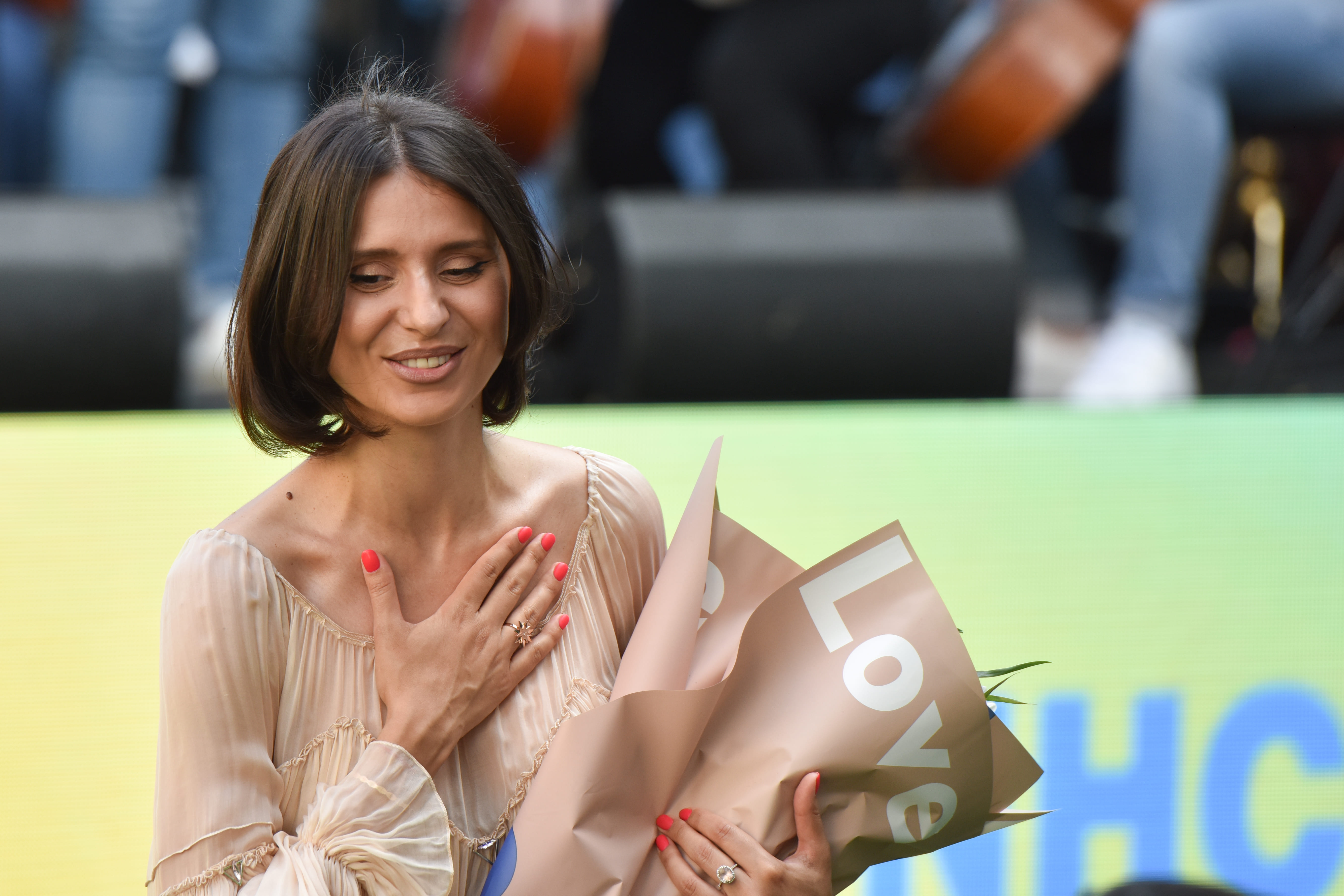 Una joven de cabello castaño que lleva puesto un vestido color beige sonríe sosteniendo un ramo de flores