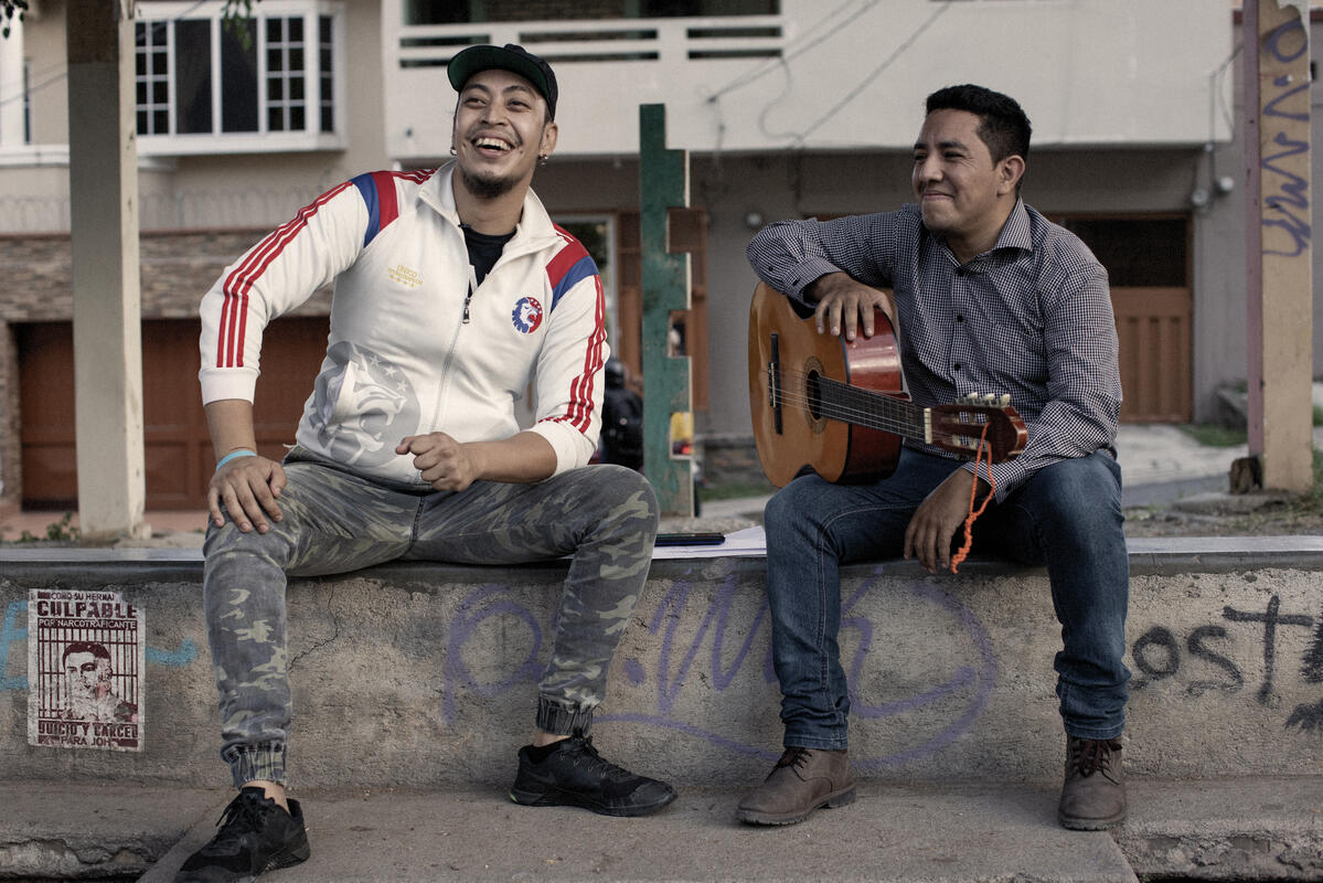 Dos hombres sentados en una barda de cemento, uno sostiene una guitarra