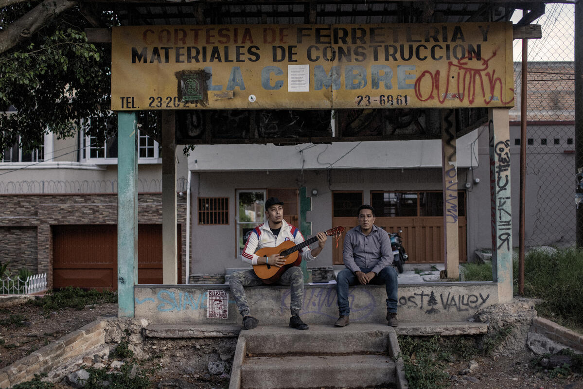 Dos hombres sentados en una barda de cemento. El de la izquierda sostiene una guitarra