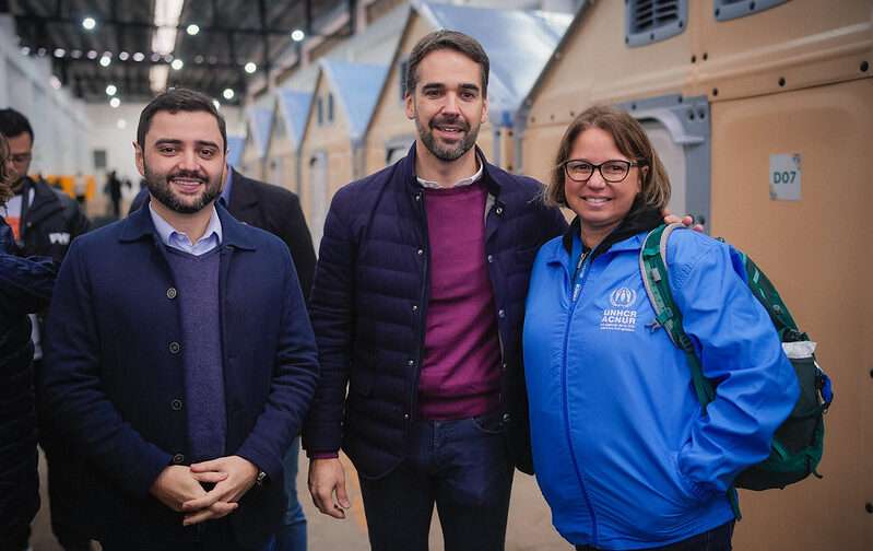 Dos hombres vestidos de negro y una mujer con chamarra azul posan para la foto frente a unas casas