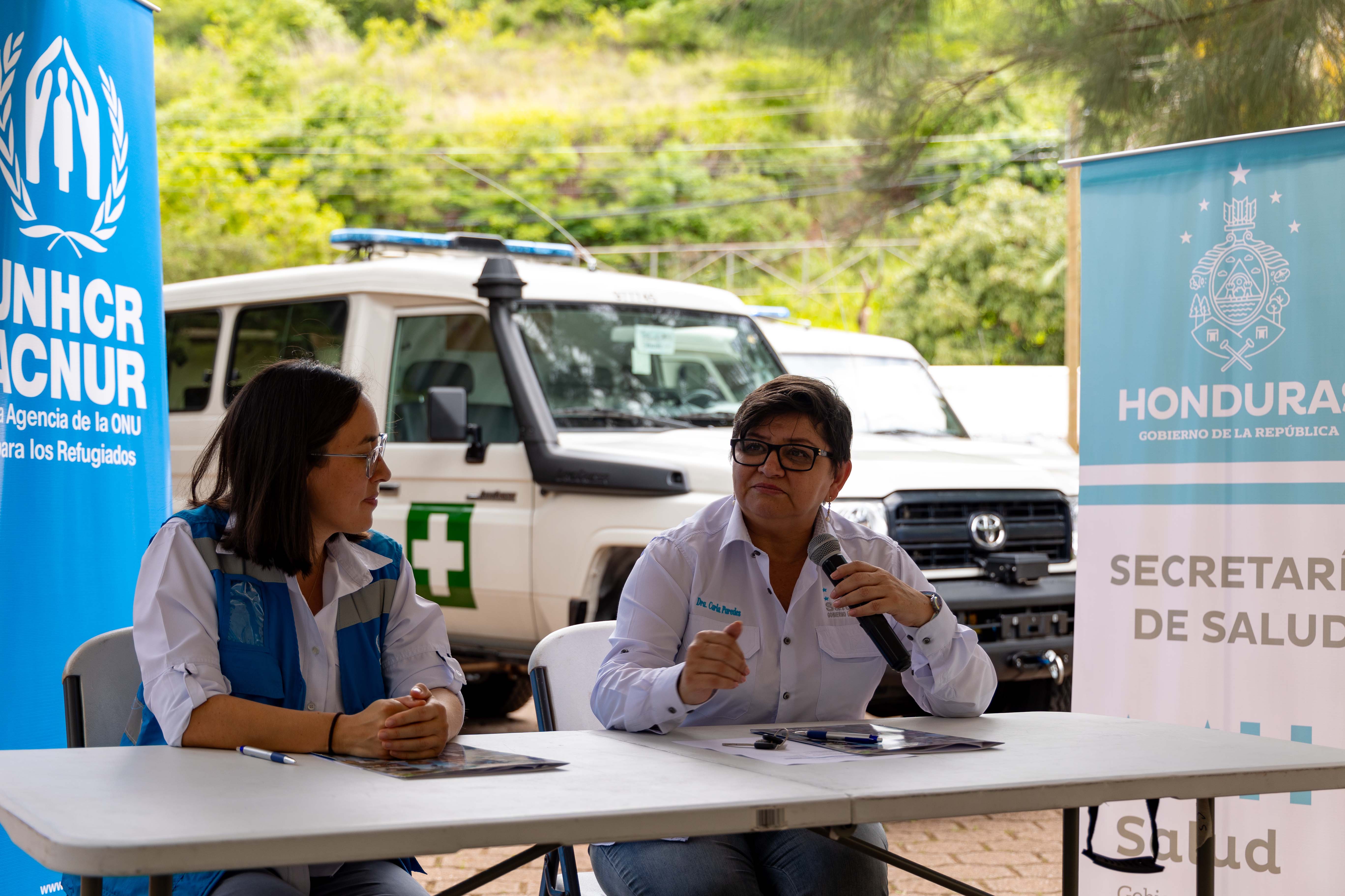 dos mujeres sentadas a una mesa. A sus lados rótulos de ACNUR y del Gobierno de Honduras. Al fondo dos ambulancias todo terreno.