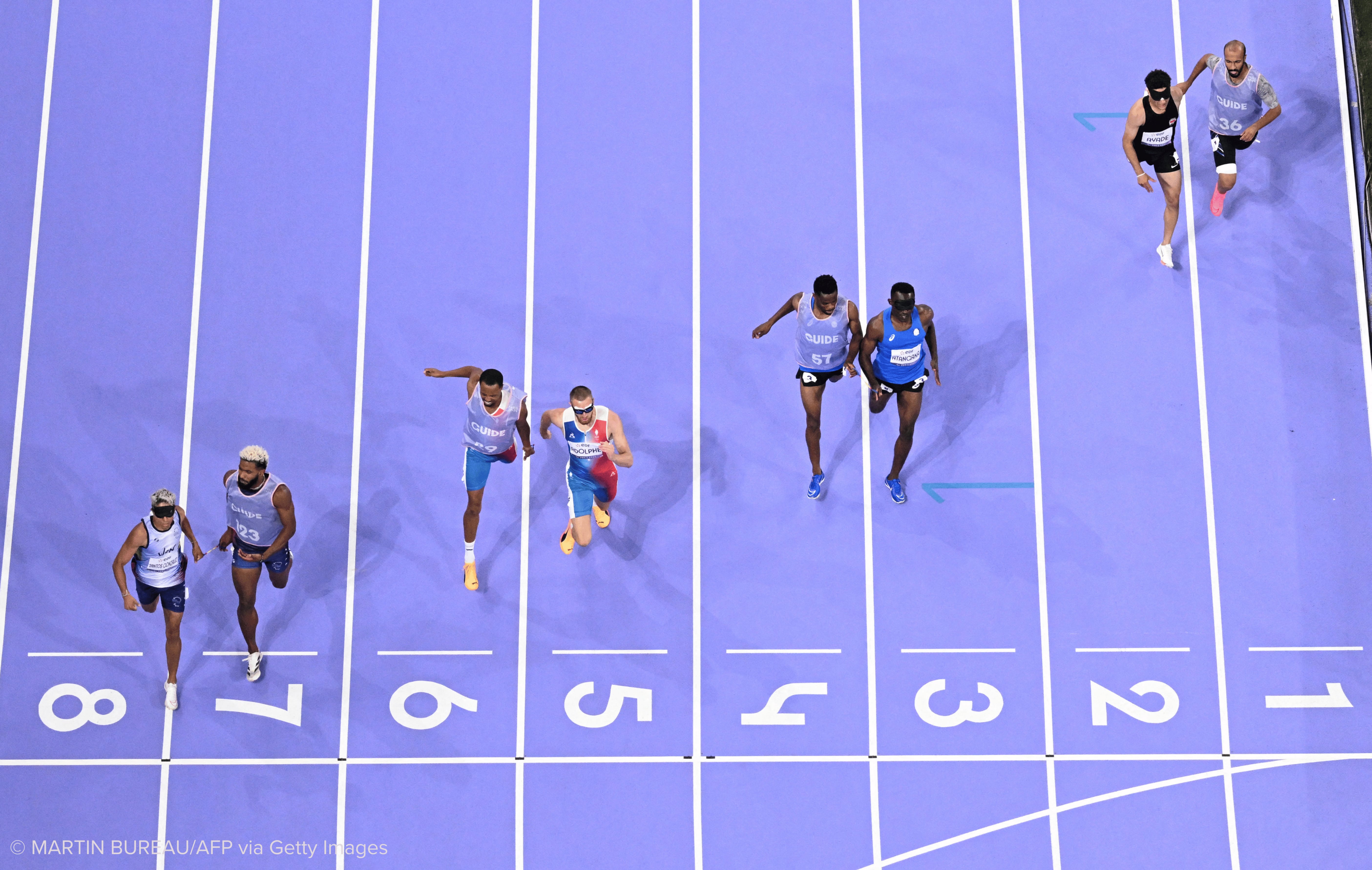 Vista aérea de cuatro parejas de corredores acercándose a la meta de una pista de atletismo de color morado con número y líneas blancas