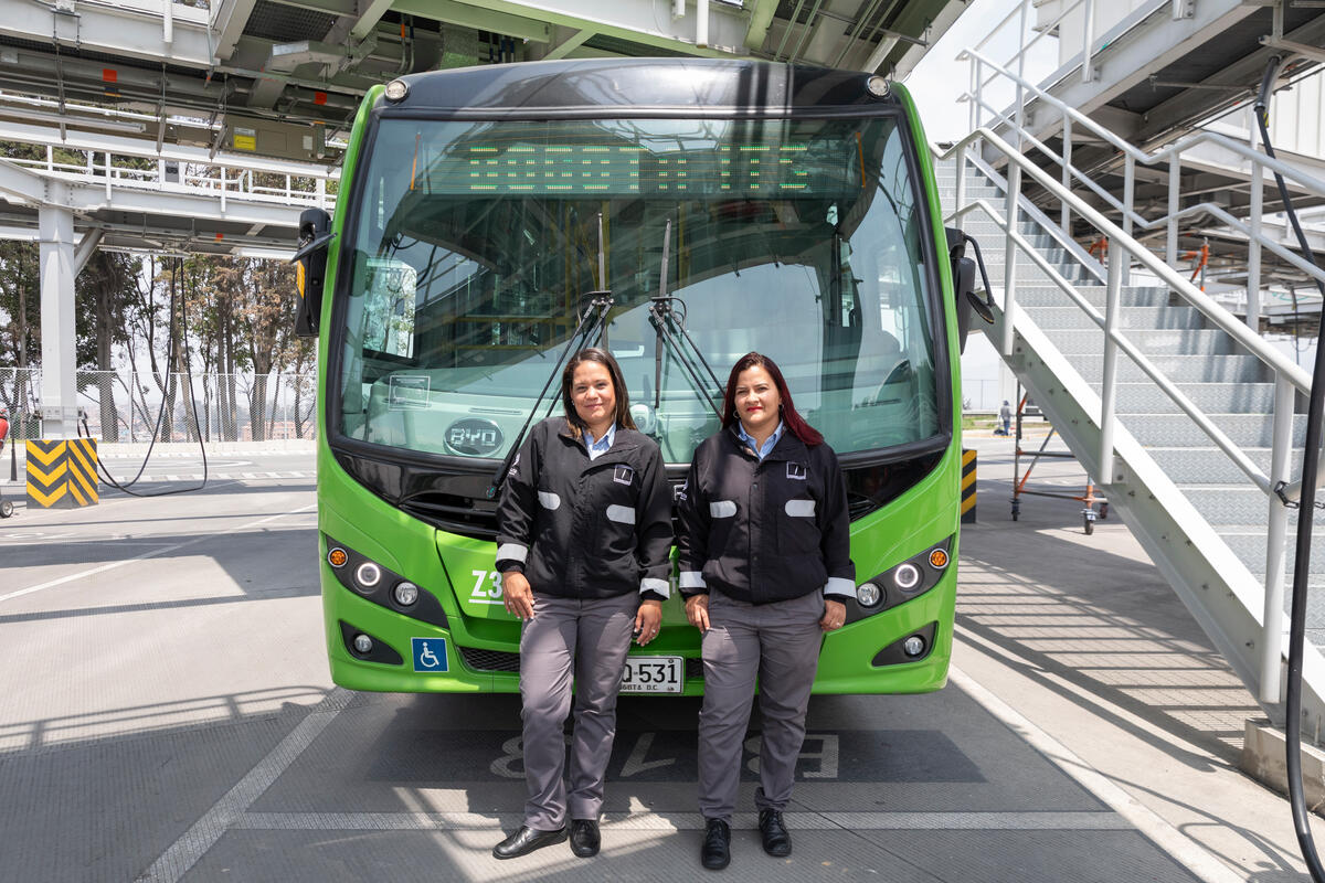 Dos mujeres con uniforme de chamarra negra y pantalón gris formal frente a un autobús de color verde