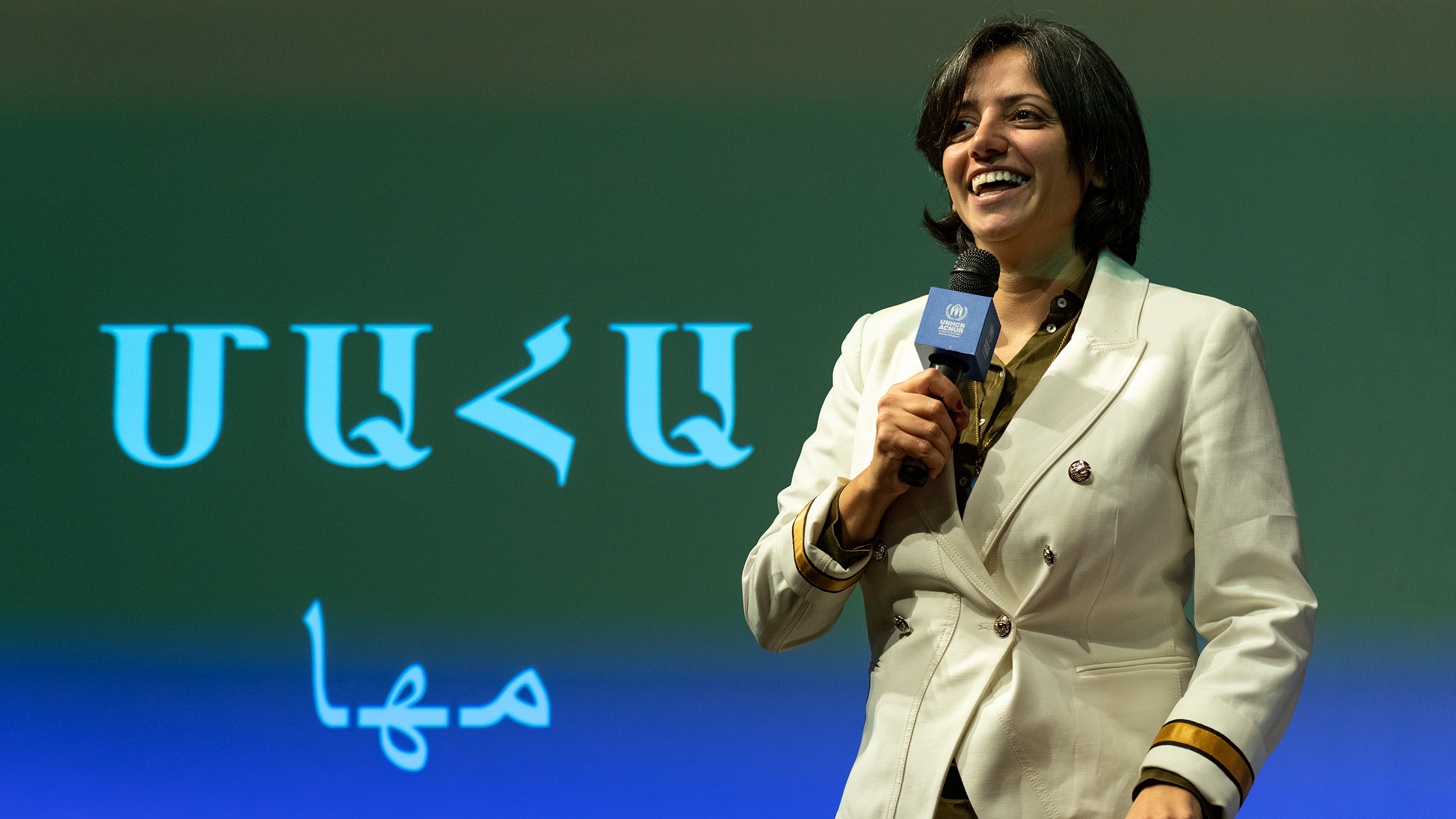 Una mujer de cabello corto oscuro, sostiene un micrófono con logotipo de ACNUR