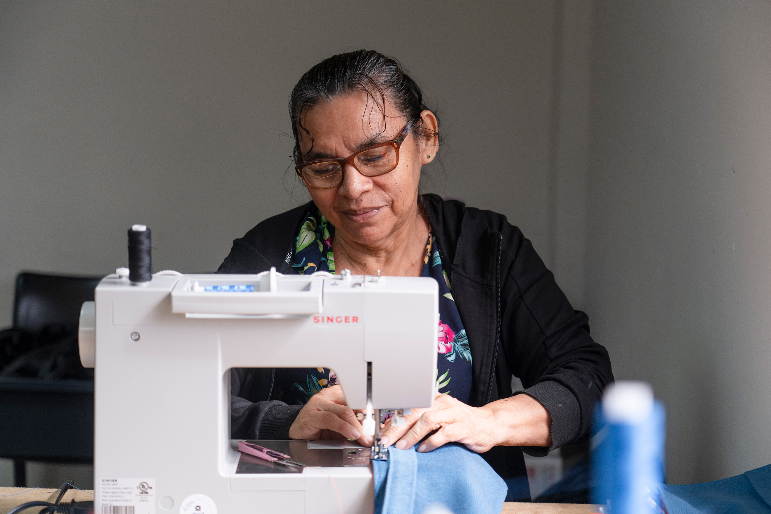 Una mujer que usa anteojos y viste suéter negro, trabaja con tela e hilo celestes frente a una máquina de coser