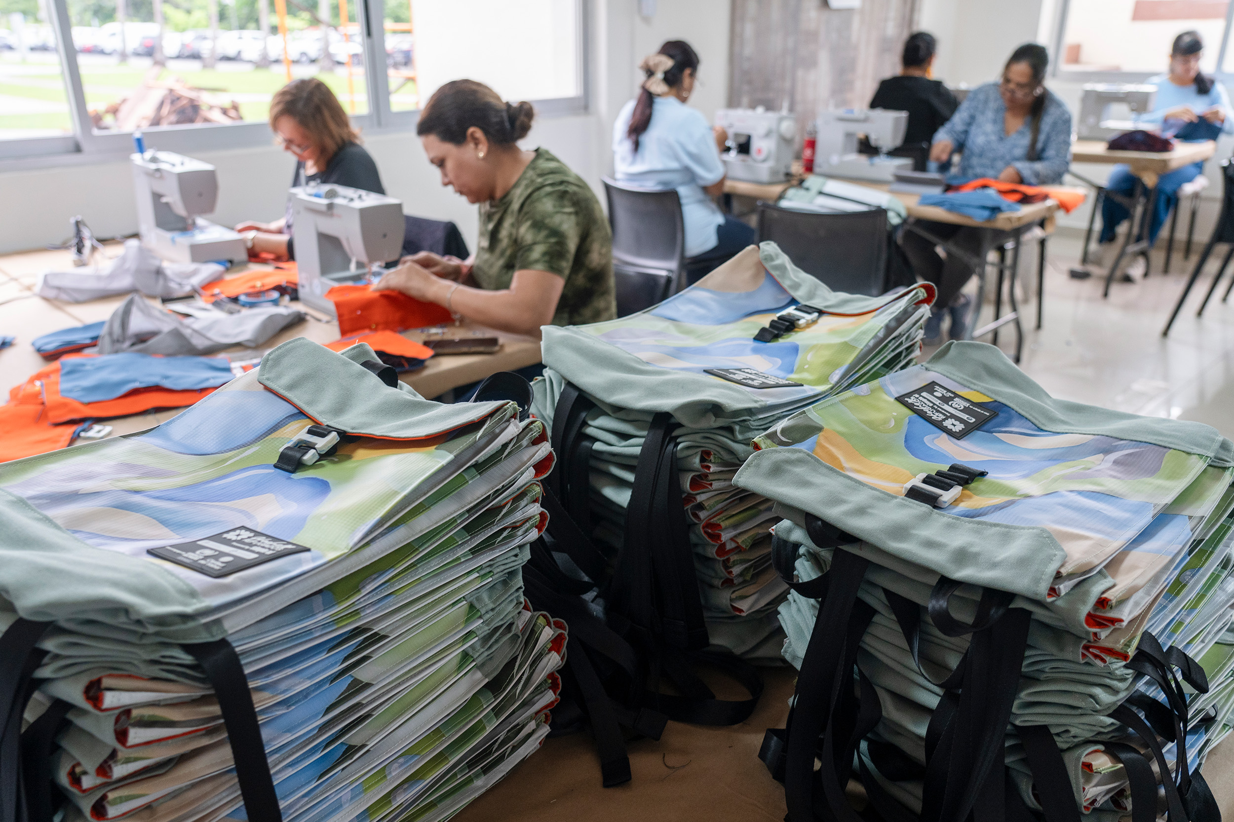 Mochilas apiladas sobre una mesa, al fondo mujeres trabajando en máquinas de coser