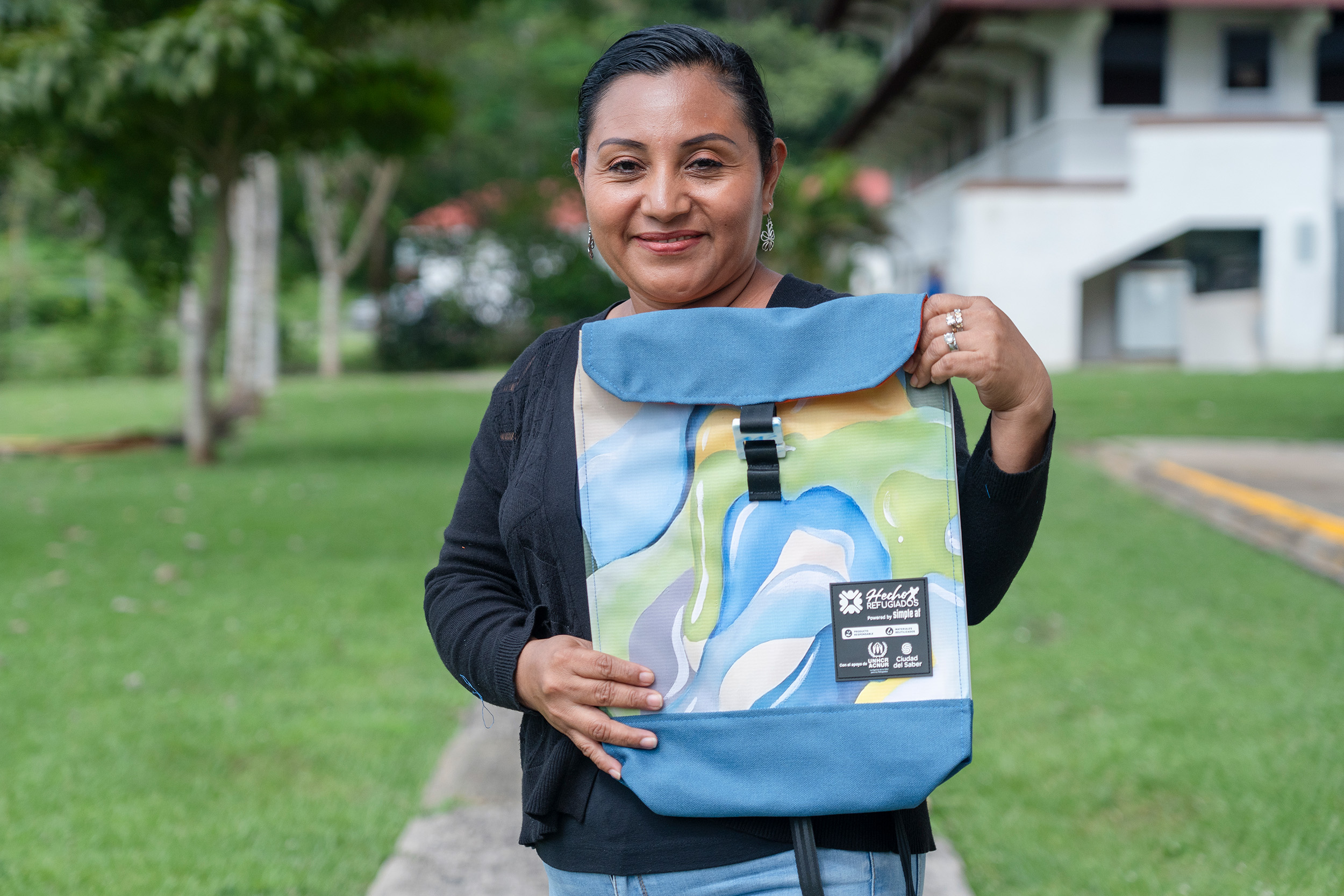Una mujer sonriente sostiene una mochila de colores en sus manos. Está parada en un patio con césped verde y árboles al fondo
