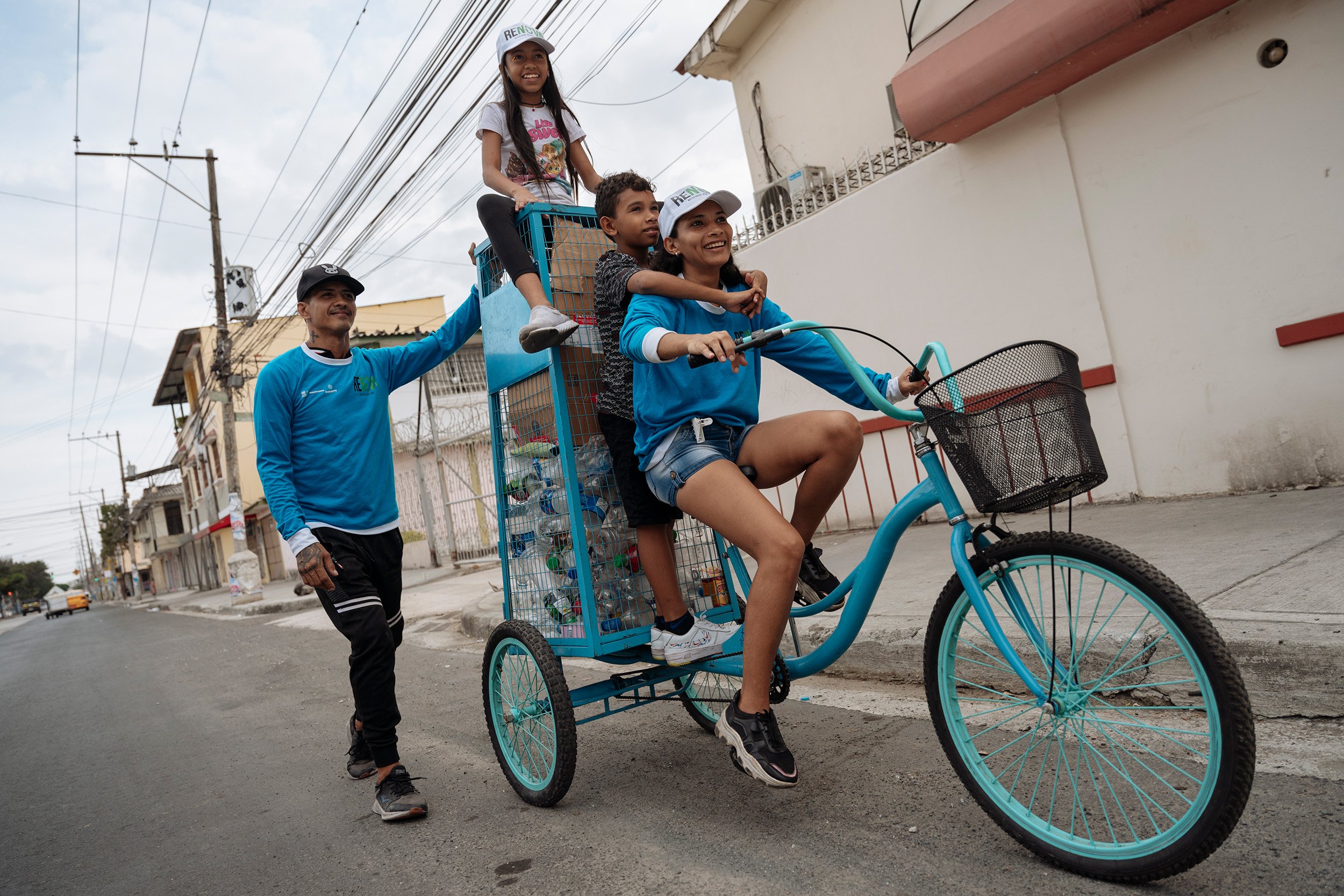 Una mujer pedalea un triciclo y carga a un niño y a una niña atrás. Un hombre los acompaña de pie sobre la calle