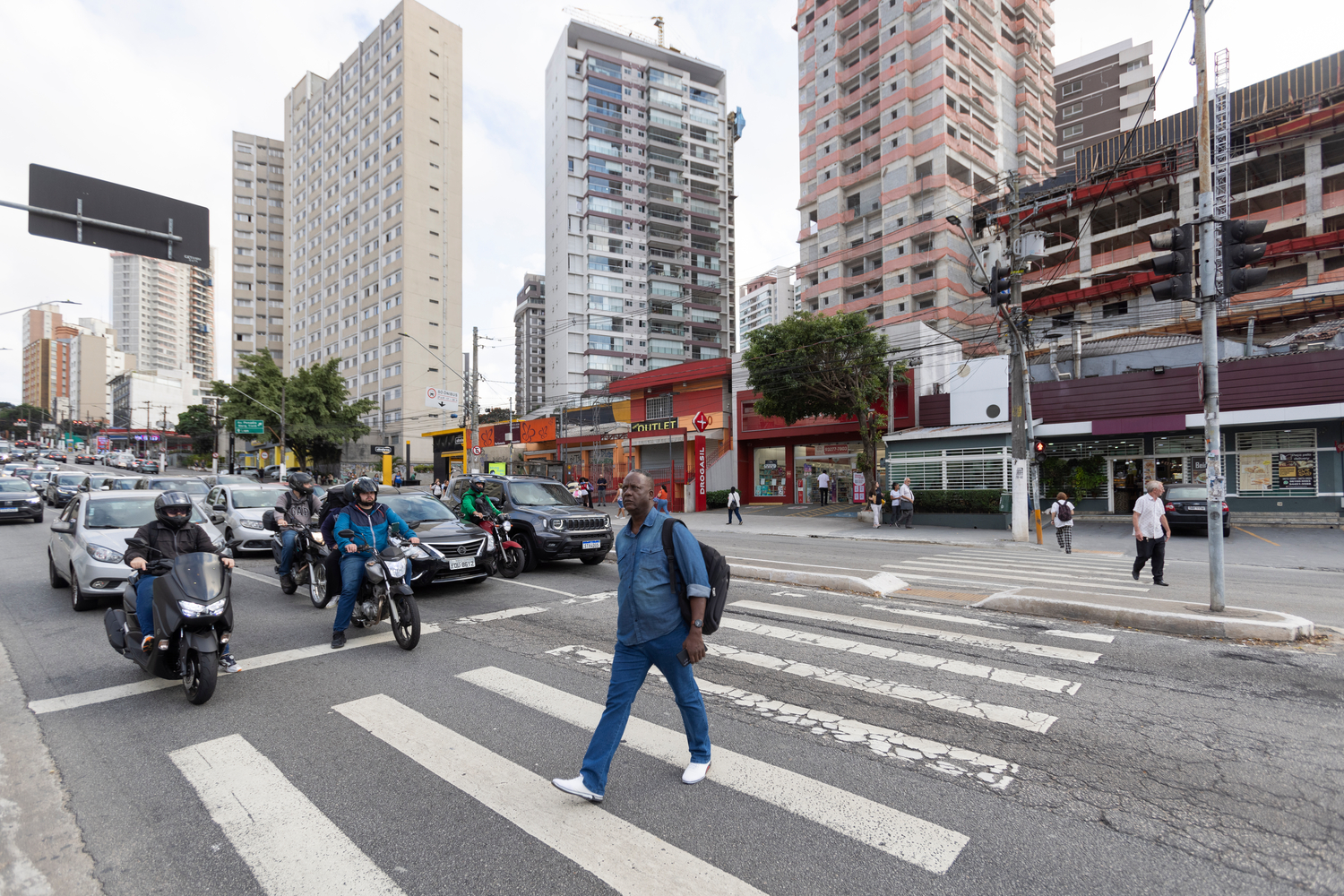 Un hombre vestido de camisa y pantalón de mezclilla lleva una mochila negra sobre su hombro izquierdo, mientras camina sobre las líneas "zebra" de una avenida transitada