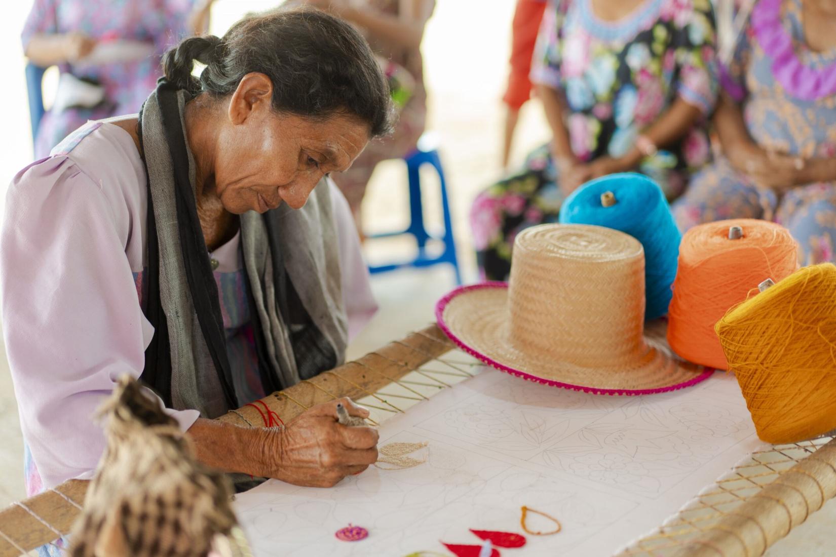 Una mujer mayor borda en una mesa al lado de hilos de colores